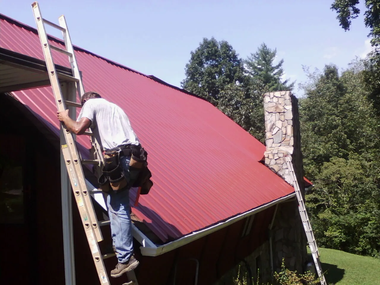 Roofer on a ladder beside a red metal roof and stone chimney — Soffit & Fascia Repair by Roofing Brothers in Willow City